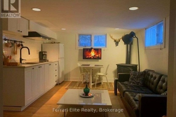 Basement living area with a small kitchen, dining table, black sofa, wood stove, and a television. - Bsmt - 66 Richmond Street, Richmond Hill