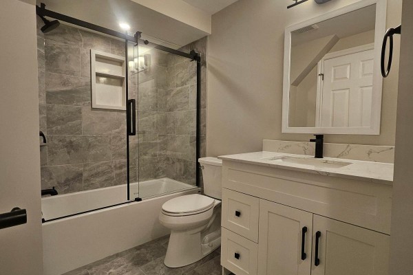 Basement bathroom with bathtub, glass shower door, toilet, white vanity, and grey tiled walls. - Bsmt - 79 Mantz Crescent, Whitby