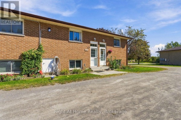 Exterior view of a brick apartment building with two entrances, gravel driveway, and grassy area. - C - 103 Rita Crescent, Kawartha Lakes