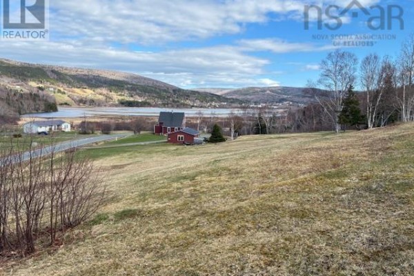 View of undeveloped grassy lot with distant red buildings, fields, water, and rolling hills under a blue sky with clouds. - Cabot Trail, Scotch Hill, Margaree Harbour