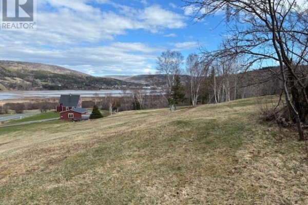 Sloping grassy lot with bare trees and birch trees, overlooking a water body and distant buildings nestled in hills. - Cabot Trail, Scotch Hill, Margaree Harbour