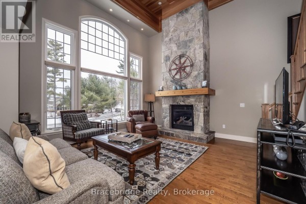 Living room with a large stone fireplace, vaulted ceiling, and floor-to-ceiling windows overlooking a snowy landscape. - Carling 10 W3 - 3876 Muskoka Rd 118 Highway W, Muskoka Lakes