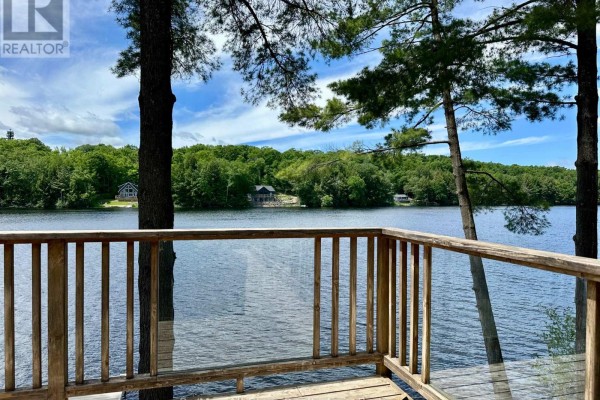 View from the wooden deck of a calm lake surrounded by green trees and distant cottages. - Cottage # 9 - 1006 Youngs Road, Muskoka Lakes