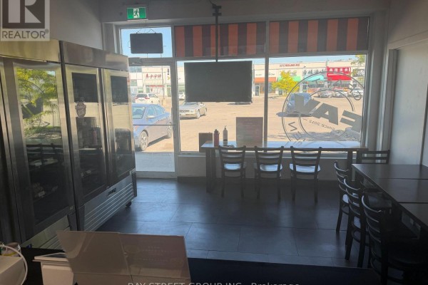 Interior view of a commercial space with stainless steel refrigerator, seating area with tables and chairs, and large front windows. - E107 - 3262 Midland Avenue, Toronto