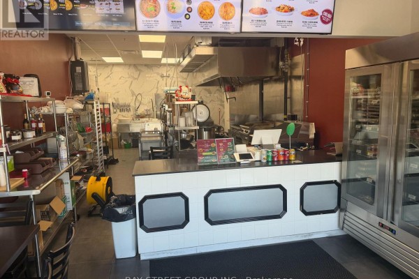 Counter area with tiled front, menu displays on wall, stainless steel shelving, and a commercial kitchen in background. - E107 - 3262 Midland Avenue, Toronto