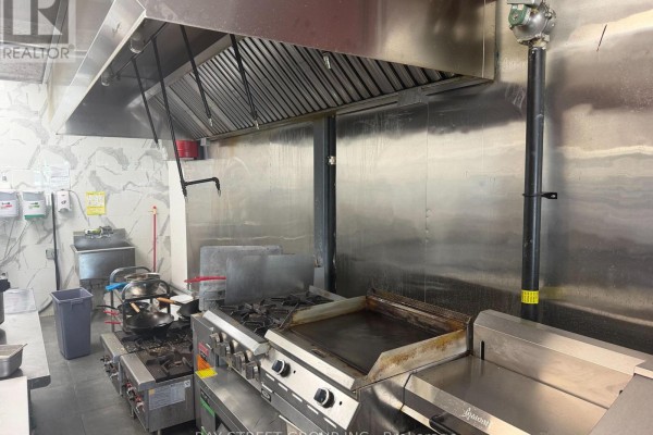 Commercial kitchen with stainless steel appliances, including a gas range and griddle, under a large exhaust hood. - E107 - 3262 Midland Avenue, Toronto