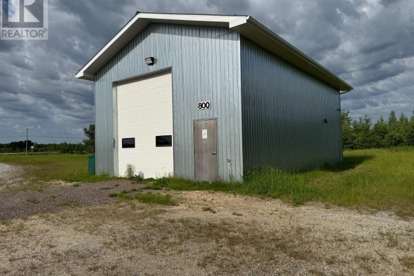 Side exterior of a metal commercial building with a roll-up garage door, a single door, and a cell tower in the distance. - Fauquier-strickland - 800 Cloutier Road, Kapuskasing