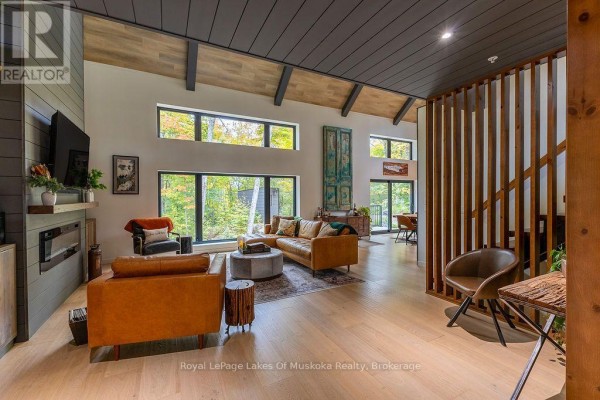 Living room with a leather sectional, fireplace, and large windows overlooking autumn trees. - Lot 18 - 113 Gryffin Bluffs Lane, Huntsville