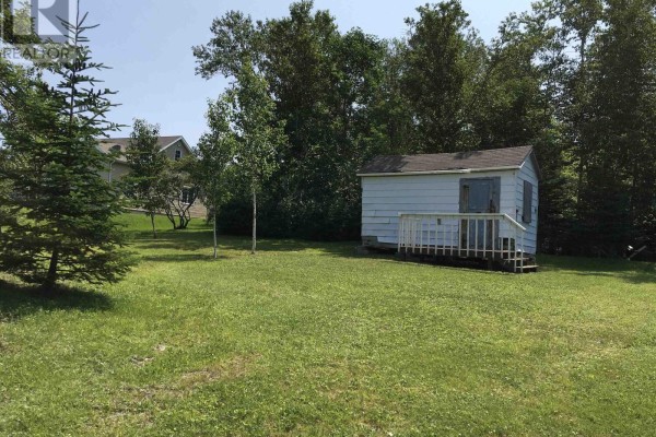 A white shed is set back in a grassy yard with trees and a house in the distance. - Lot 2 Cabot Trail, La Prairie