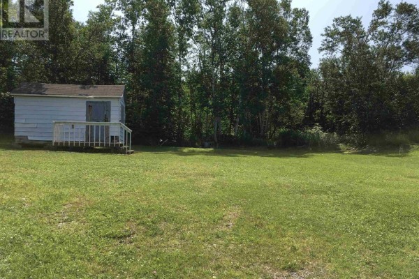 A white shed with a porch sits in a wide, flat grassy area with trees bordering the property. - Lot 2 Cabot Trail, La Prairie