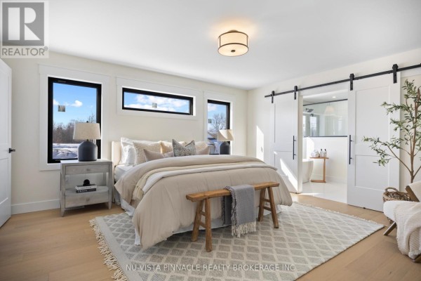A spacious primary bedroom with a king-size bed, sliding barn doors to the ensuite, and large windows. - Lot 20 Harvest Lane, Thames Centre