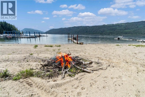 A bonfire burns on a sandy beach with a lake and distant mountains in the background. - Lot 4 Queest N. Bay, Sicamous