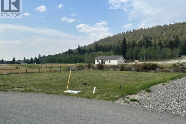 View of the lot with gravel road, grass area, and distant house among trees. - Lot 8-110 Forest Ridge Road, 100 Mile House