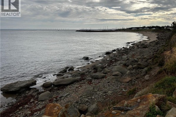 View of a rocky coastline and calm ocean with a distant bridge and town under a cloudy sky. - Lot Route 955, Murray Corner