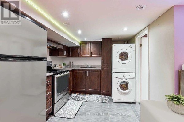 A galley kitchen with dark wood cabinets, stainless steel appliances, and a stacked washer/dryer. - Lower - 10 Muriel Avenue, Toronto