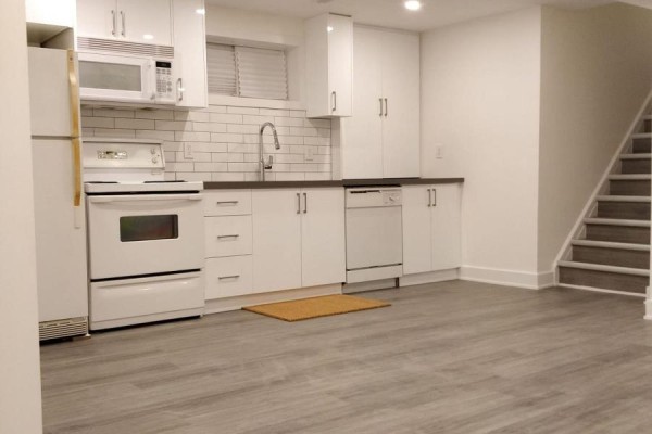 Kitchenette with white cabinets, white appliances, and stairs leading up to the right. - Lower - 1185 Woodbine Avenue, Toronto