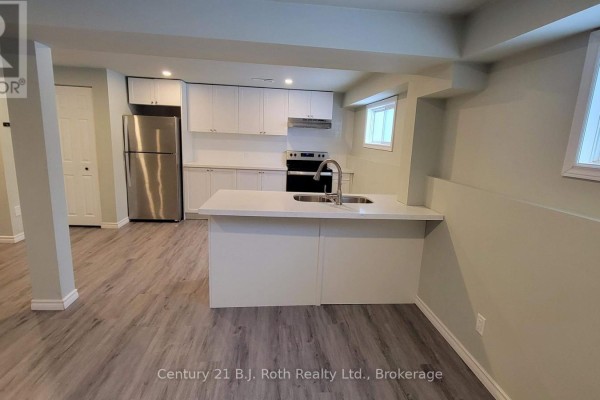Open-concept basement with a kitchen, island, stainless steel refrigerator, and light grey flooring. - Lower - 12 Shannon Street, Orillia