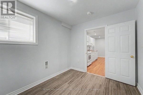 A room with a window, light gray walls, and light wood floors, opening to a kitchen area. - Lower - 148 Livingstone Street E, Barrie