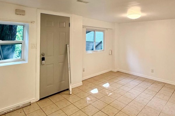 Room with beige tiled floors, white walls, a grey door, and two windows with greenery outside. - Lower - 191 Coxwell Avenue, Toronto