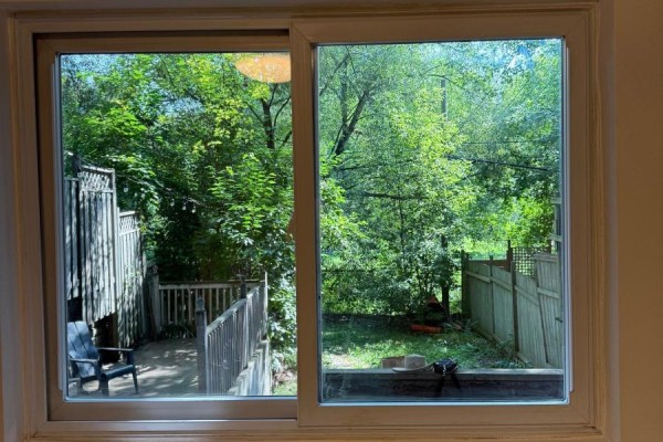 View through a sliding window of a backyard with a wooden deck, fence, and lush trees. - Lower - 191 Coxwell Avenue, Toronto