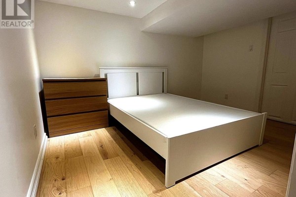 A bedroom with a white bed frame, white headboard, and a wooden dresser on light wood floors. - Lower - 25 Mann Avenue, Toronto