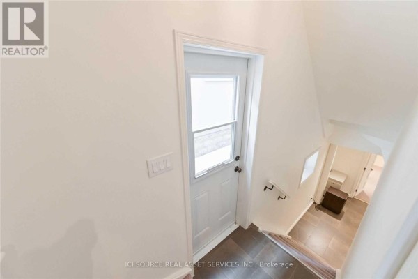 Interior view of a hallway with a white door, staircase with handrail, and tiled flooring. - Lower - 25 Mann Avenue, Toronto