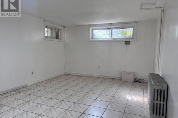 Empty room with tiled floor, white walls, two windows, and a large cast iron radiator. - Lower - 415 Delaware Avenue, Toronto