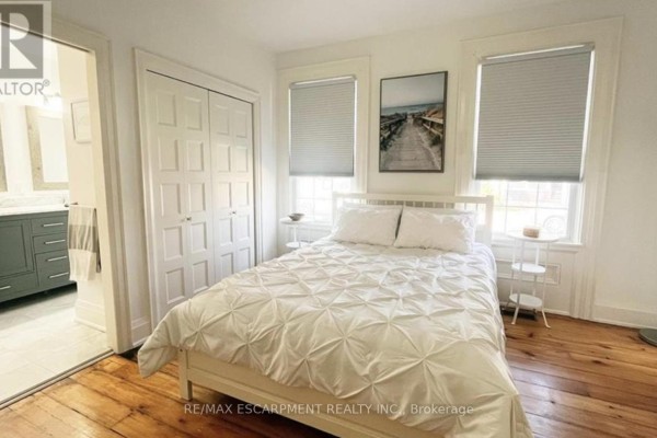 Bedroom with a white bed, white closet doors, and a painting above the bed. - Main - 32 Rodman Street, St  Catharines
