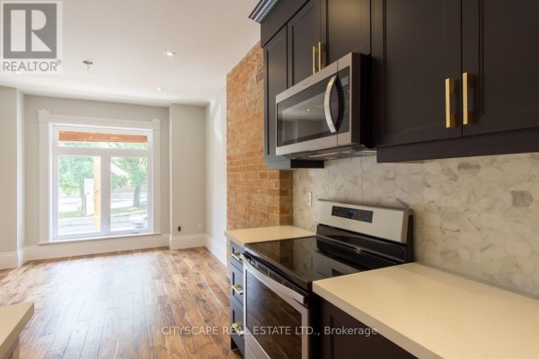 Kitchen with dark cabinets, stainless steel microwave and stove, white quartz countertops, and exposed brick wall. - Main - 936 Dundas Street E, Toronto