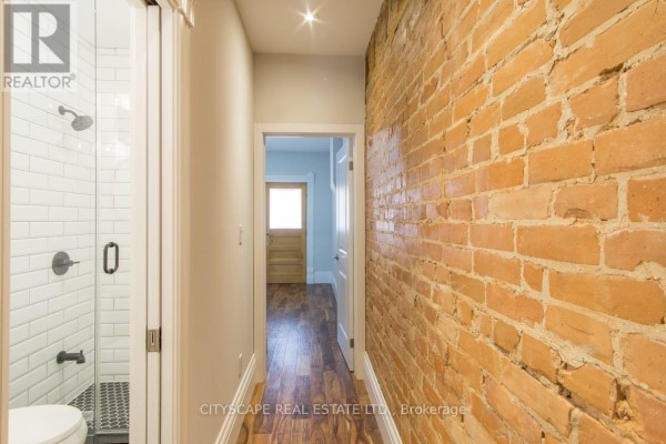 Hallway with exposed brick wall, hardwood floors, and a shower stall with white subway tiles on the left. - Main - 936 Dundas Street E, Toronto