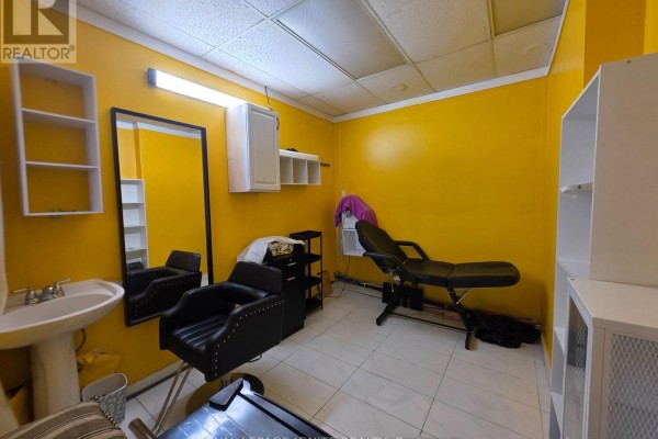 A smaller room with bright yellow walls, a black styling chair, a massage table, and a sink. - Main Floor (front Side) - 2426 Kingston Road, Toronto