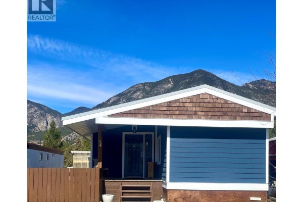 Backyard with a metal bench under an arbor, wooden fence, and pine trees with mountain view. - Pad #14 - 5174 Lambert Road, Invermere