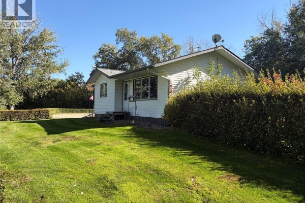 Exterior view of the white bungalow showing the side entrance and surrounding green lawn and trees. - Peterson Acreage, Big Quill Rm No  308