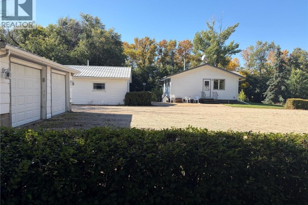 View of the white bungalow and detached garage from behind a hedge with a gravel driveway. - Peterson Acreage, Big Quill Rm No  308