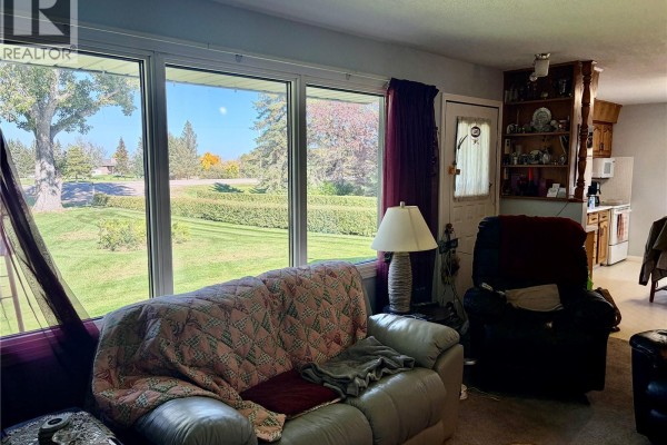 Living room with large windows overlooking a lawn, a couch, a recliner, and a partial view of the kitchen. - Peterson Acreage, Big Quill Rm No  308