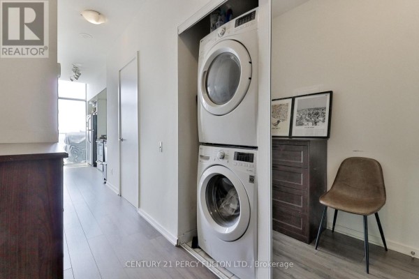 Stacked white washer and dryer in a closet with a dark dresser and chair. - Ph3505 - 105 The Queensway Avenue, Toronto