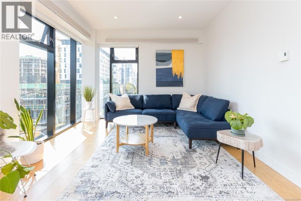 Living room with a blue sectional sofa, marble coffee table, patterned rug, and large windows with city views. - S404 1411 Cook St, Victoria