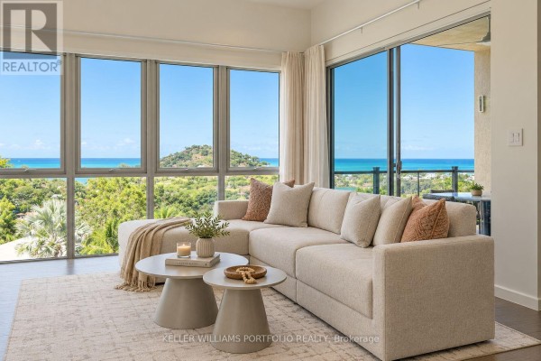 Living room with a large sectional sofa and panoramic windows offering ocean and greenery views. - Sky Residence - Sugar Ridge Condos Valley Road, Antigua