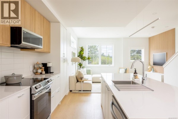 View from kitchen island towards living room with white sectional sofa and large windows. - Th6 3450 Whittier Ave, Saanich