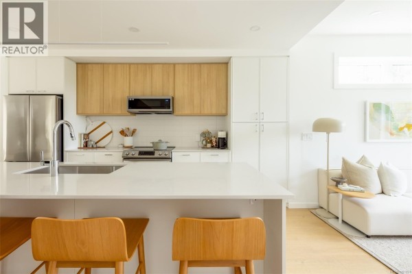Kitchen island with light wood stools, white cabinets, stainless steel appliances, and living room view. - Th6 3450 Whittier Ave, Saanich