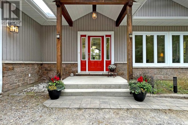 Close-up of a front entrance with a red door, covered porch, stone foundation, and potted plants. - Unit 4 - 1623 Houseys Rapids Road, Gravenhurst