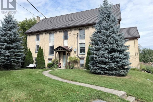 Front exterior of a multi-unit brick building with manicured lawns and large evergreen trees. - Unit 5 - 108 Queen Street N, West Grey