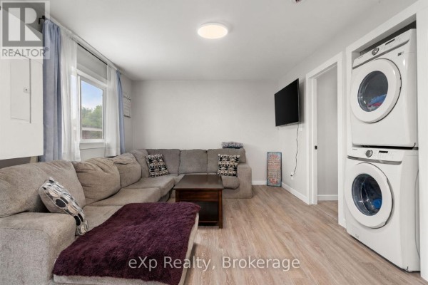 Living area with grey sectional sofa, television, and stacked white washer and dryer unit. - Unit A - 89 College Street E, Belleville