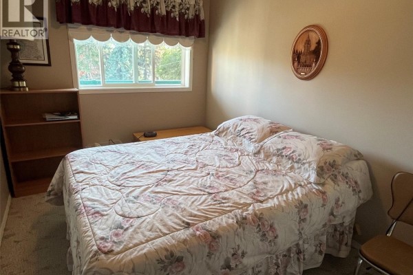 Bedroom with a double bed, floral comforter, and a window overlooking trees. - Unit306 1 26th Street E, Prince Albert