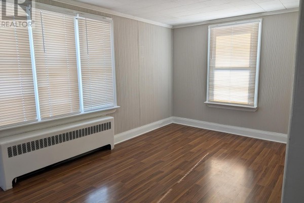 Empty room with wood-look laminate flooring, pale grey textured walls, and a white radiator under a window. - Upper - 12 East 24th Street, Hamilton