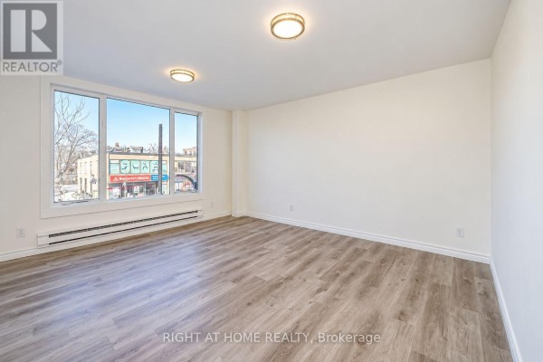 Empty living room with light wood floors and a large window overlooking Danforth Avenue. - Upper - 843 Danforth Avenue, Toronto