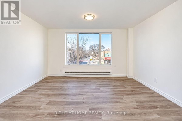 Empty room with light wood floors and a large window facing a street view. - Upper - 843 Danforth Avenue, Toronto