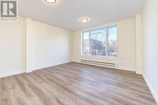Empty room with light wood floors and a large window overlooking a street scene. - Upper - 843 Danforth Avenue, Toronto