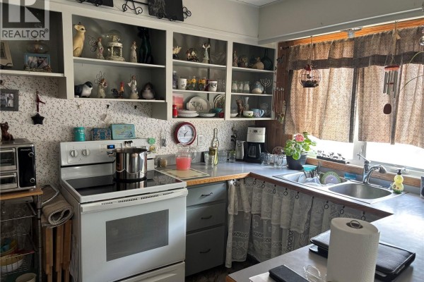A kitchen with white appliances, grey cabinets, open shelving, and floral curtains over the sink. - Welsh Acreage, Excelsior Rm No  166